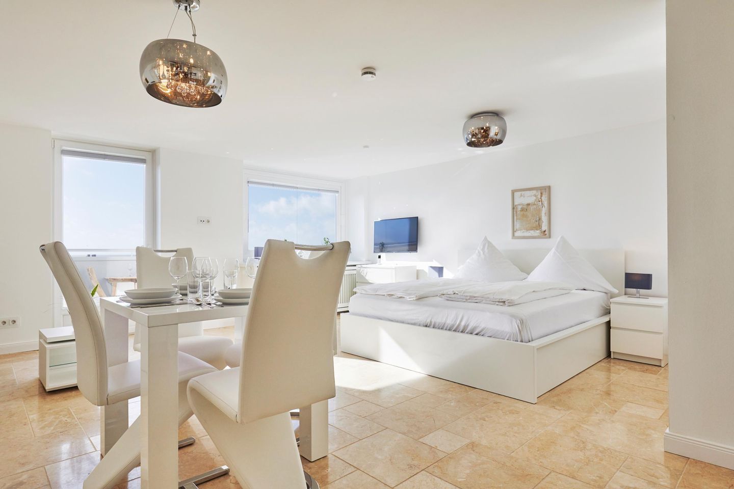 A modern bedroom with a large bed, a dining area with chairs, a TV, and a view of the sky.