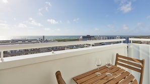 A balcony with a wooden table and two wine glasses, overlooking a coastal area with buildings and a view of the sea. - Sea view I - Modern apartment, balcony with sea view, parking space, WLAN. (Sylt)