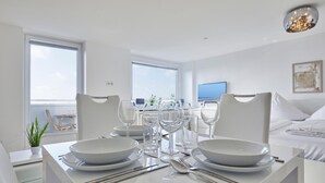 A modern dining area with a white table set for two, a sofa in the background, and a view of the sea through large windows. - Sea view I - Modern apartment, balcony with sea view, parking space, WLAN. (Sylt)
