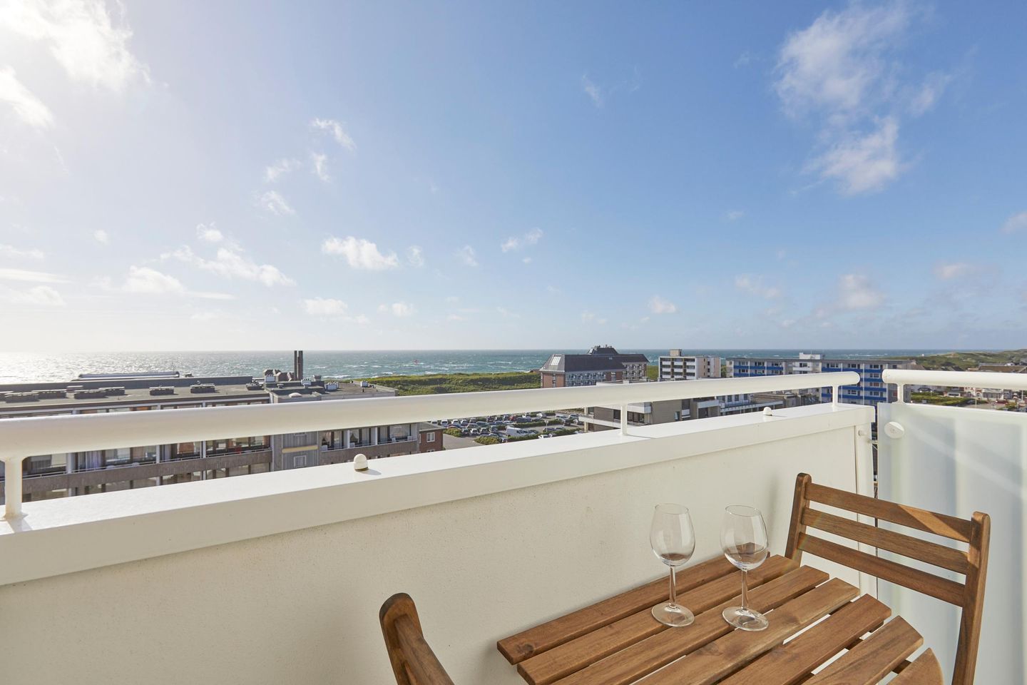 Een balkon met een houten tafel en twee wijnglazen, met uitzicht op een kustgebied met gebouwen en de zee.