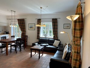Een gezellige woonkamer met een zwarte leren bank, een houten salontafel en een eethoek met een houten tafel en stoelen. Er zijn twee ramen met gordijnen, een plafondventilator en een lamp op een bijzettafeltje.