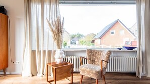 Ein Zimmer mit Holzboden, ein Stuhl mit gemustertem Sitzkissen, ein kleiner Holztisch, eine Vase mit Trockenpflanzen und Blick auf ein Backsteinhaus durch ein Fenster mit weißen Vorhängen.