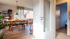 Ein modernes Wohnzimmer mit einem Holzesstisch, Stühlen, einem Bücherregal und einem großen Fenster mit Vorhängen.
