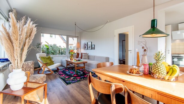 Ein modernes Wohnzimmer mit einem Holzespeisetisch, einer Couch und einem Couchtisch. Es gibt eine Küchenzeile mit einer Pendelleuchte und einer Obstschale auf dem Tisch.