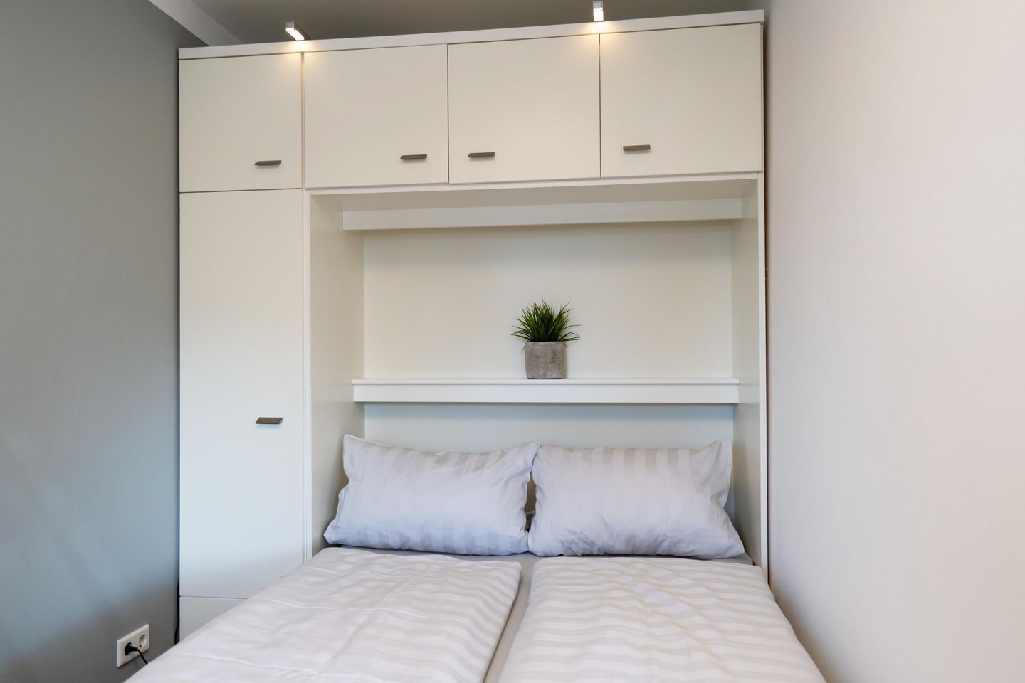 Un dormitorio con una cama, dos almohadas, una estantería con una planta y armarios blancos.