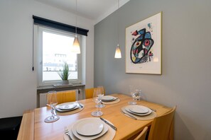 A dining area with a wooden table set for four, a window with blinds, a radiator, and a framed abstract painting on the wall.