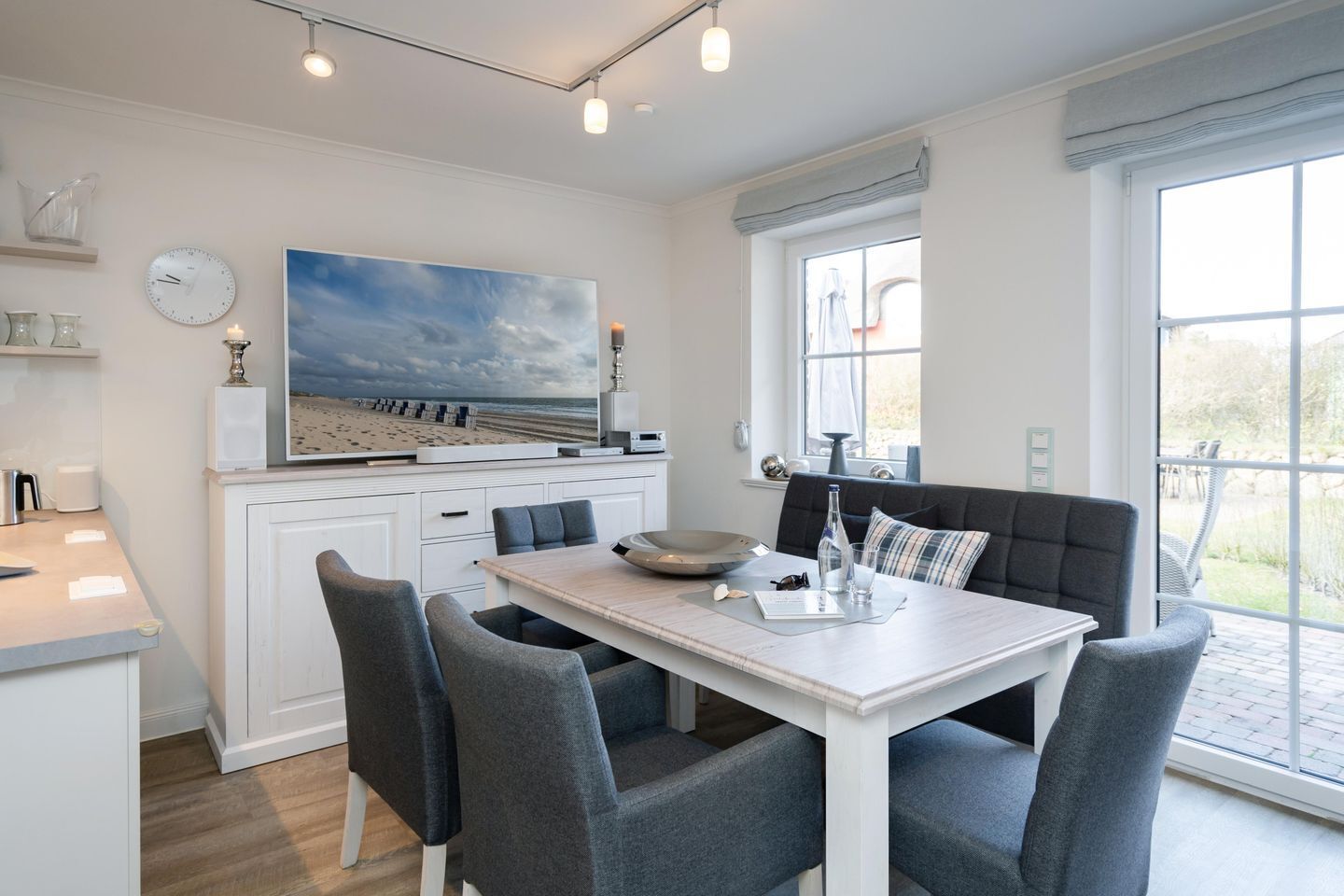 A modern kitchen with a dining area, featuring a large window, a clock, and a beach-themed artwork.