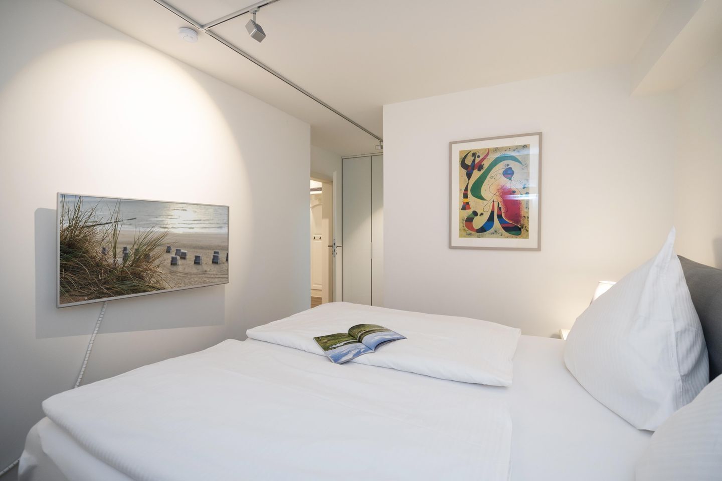A hotel room with a bed, a painting of a beach, and a colorful abstract painting on the wall.