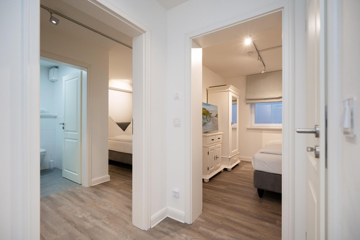A modern bedroom with a bed, a dresser, and a nightstand. There is a bathroom with a toilet and a shower visible through an open door.