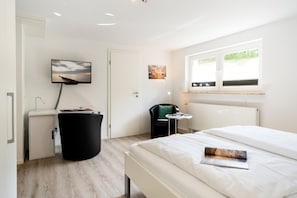 Een moderne hotelkamer met een bed, een bureau met stoel, een klein tafeltje, een prullenbak en een raam met uitzicht op groen.
