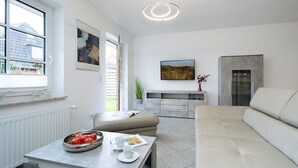 A modern living room with a sofa, a dining table with a bowl of fruit, and a kitchen area with a fridge and a TV. - Albatros Ground Floor - Modern 3-room apartment for 4 people with a terrace, garden, and parking space (Sylt)