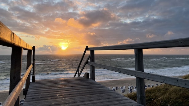 A wooden boardwalk leading to a beach with a view of the ocean and a sunset. - Albatros Ground Floor - Modern 3-room apartment for 4 people with a terrace, garden, and parking space (Sylt)