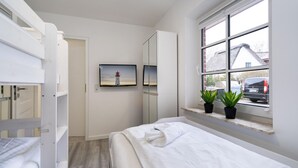 A bedroom with a bed, a wardrobe, a mirror, a window with plants, and a picture of a lighthouse. - Albatros Ground Floor - Modern 3-room apartment for 4 people with a terrace, garden, and parking space (Sylt)