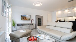A modern living room with a sofa, ottoman, and a table with apples and cups. There is a kitchen area with cabinets and a countertop. - Albatros Ground Floor - Modern 3-room apartment for 4 people with a terrace, garden, and parking space (Sylt)