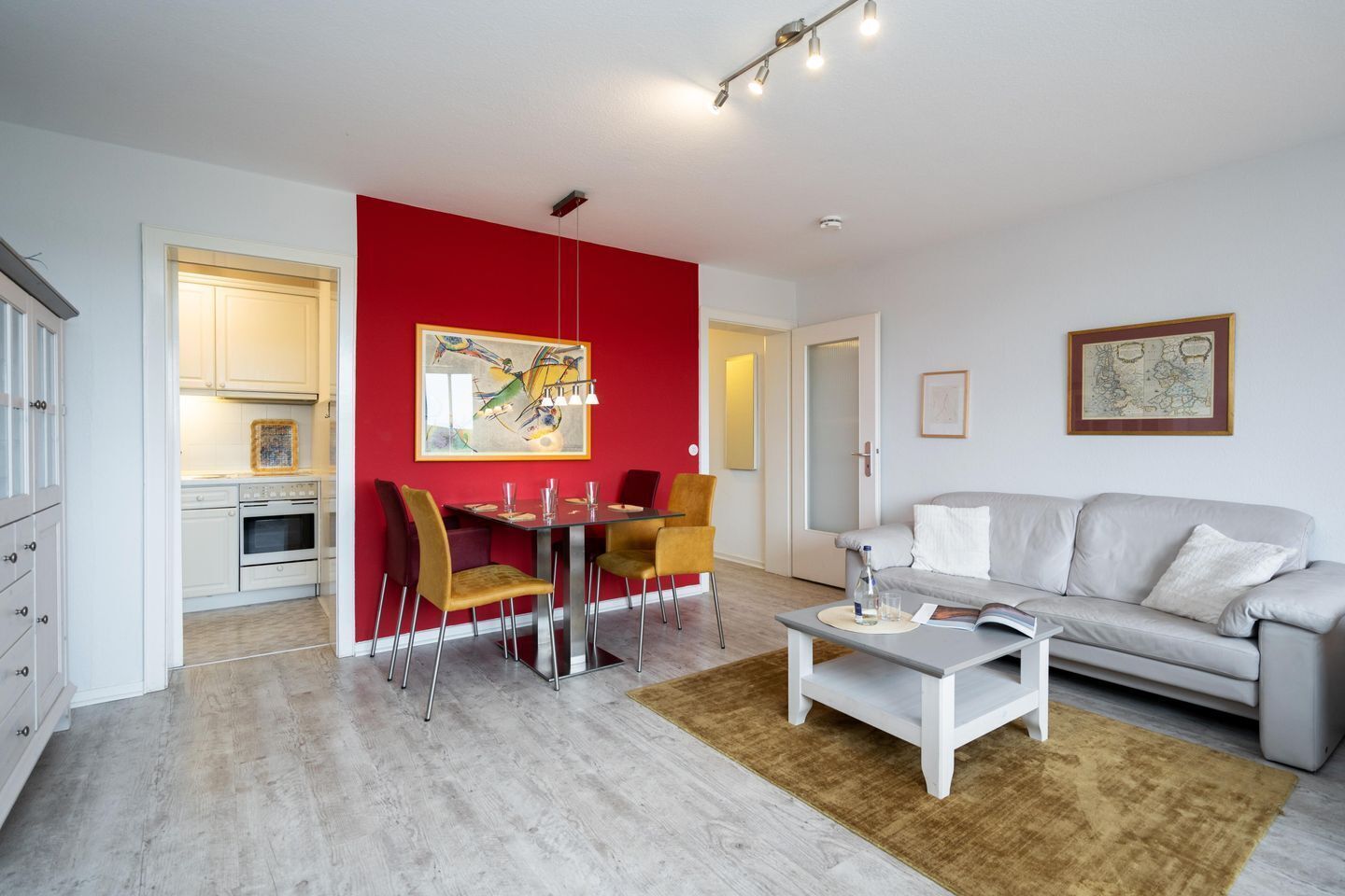 Un salon moderne avec un mur d’accent rouge, un coin repas avec des chaises jaunes, un canapé blanc et une cuisine en arrière-plan.