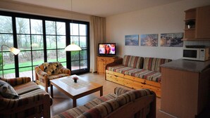 A living room with a sofa, armchairs, a coffee table, and a TV. There are large windows, a lamp, and a microwave on the counter. - Vacation apartment Warfstr. 3/3 - Wohnpark Werdum (Werdum)