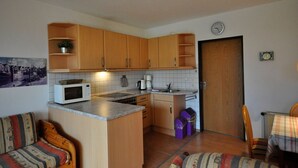 A compact kitchen with wooden cabinets, a microwave, a sink, and a dining area with a plaid tablecloth. - Vacation apartment Warfstr. 3/3 - Wohnpark Werdum (Werdum)
