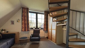 Un espacio habitable compacto con un sofá, una silla, una mesita, una ventana con cortinas y una escalera de madera.