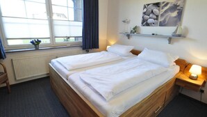 A double bed with white linens, a bedside table with a lamp, a window with a view, and a wall-mounted shelf with decorative items. - Vacation apartment Warfstr. 5/3 - Wohnpark Werdum (Werdum)