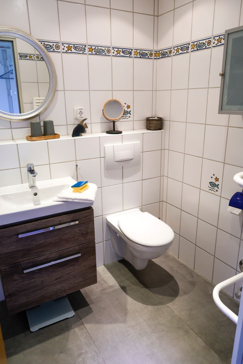 Un baño con azulejos blancos, un mueble de tocador de madera, un inodoro, un espejo y un estante pequeño con artículos de aseo.