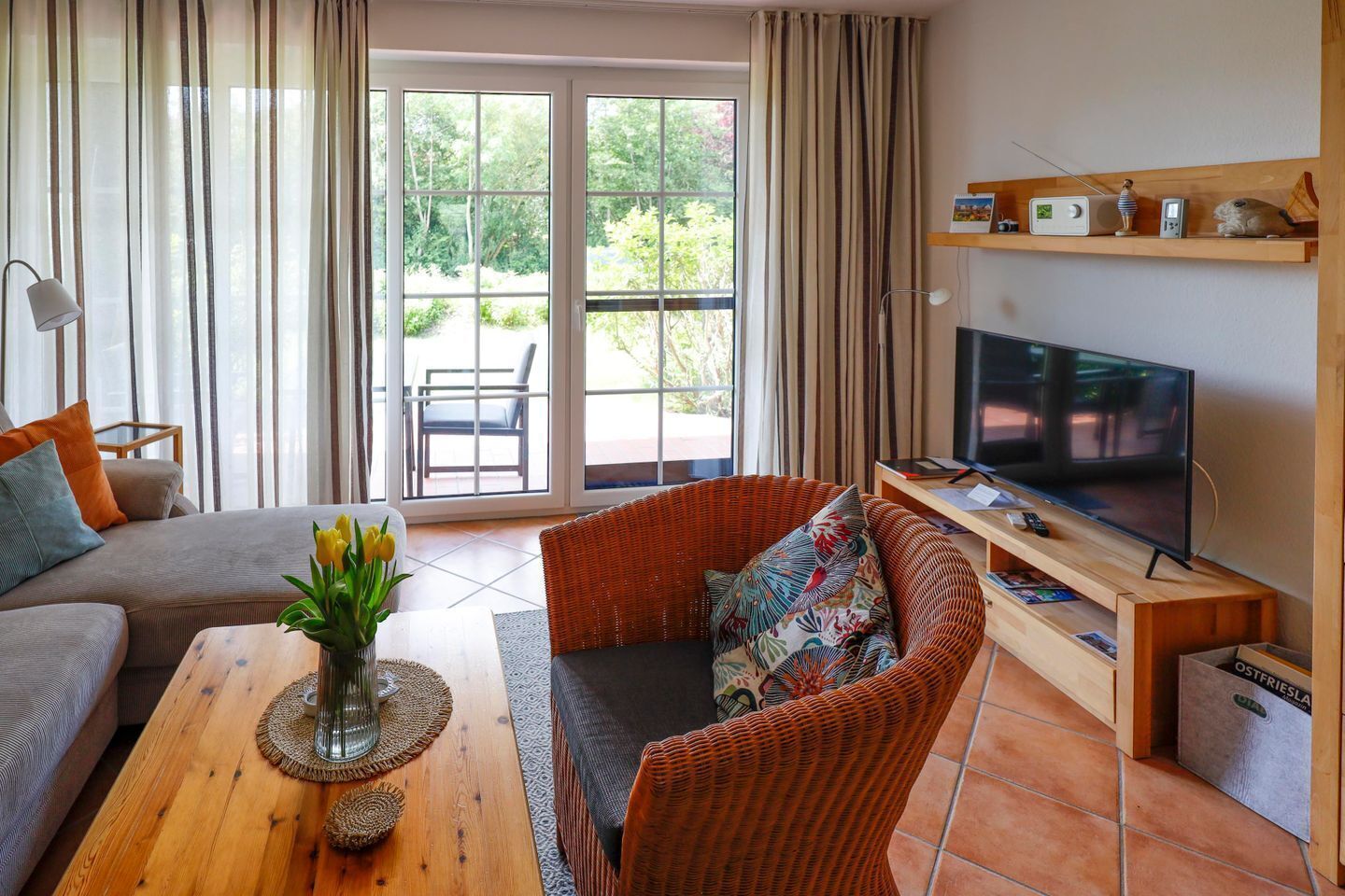 A living room with a sofa, a wicker chair, a coffee table with a vase of flowers, a television, and a shelf with decorative items.