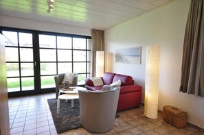 Un salón con un sofá rojo, un sillón gris y una pequeña mesa redonda. Se ve un ventanal con cortinas, una lámpara de pie y un cuadro en la pared.