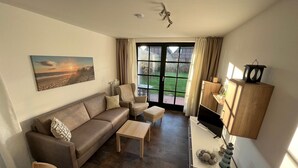 A modern living room with a sofa, armchair, and coffee table. There is a TV on a stand and a cabinet with decorative items. A large window offers a view of the outdoors. - "Friesengeist" Werdum - Werdum residential park (Werdum)