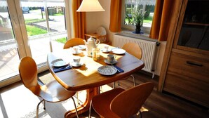 Un coin repas avec une table en bois dressée pour quatre personnes, des chaises, un radiateur, une fenêtre avec des rideaux et une lampe suspendue.