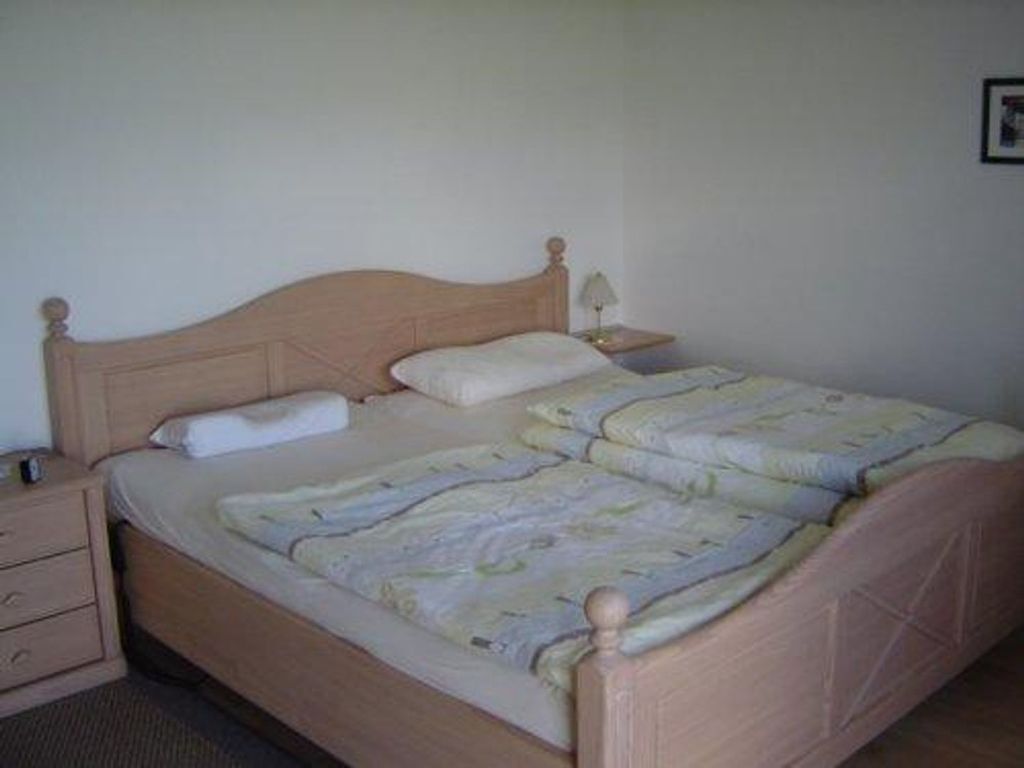 Une chambre avec un lit simple, une tête de lit en bois, une table de chevet avec une lampe et un petit tiroir.