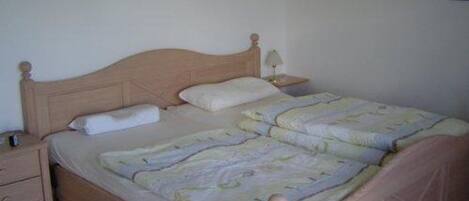 Une chambre avec un lit simple, une tête de lit en bois, une table de chevet avec une lampe et un petit tiroir.