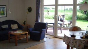 A living room with a sofa, armchair, and coffee table, a dining table with chairs, and a view of a garden through a glass door.
