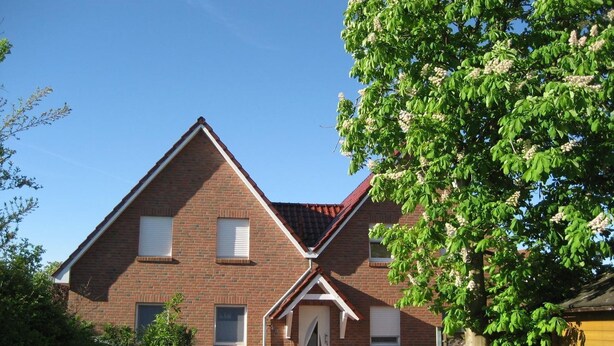 Een twee verdiepingen tellend bakstenen huis met een rode dakbedekking, een voordeur en een kleine voortuin met een bloempot.