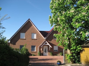 A two-story brick house with a red-tiled roof, a front door, and a small front yard with a potted plant.