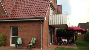 Een bakstenen huis met een rode dakbedekking, een overdekte patio met twee gestreepte stoelen, een tafel en een rode parasol.