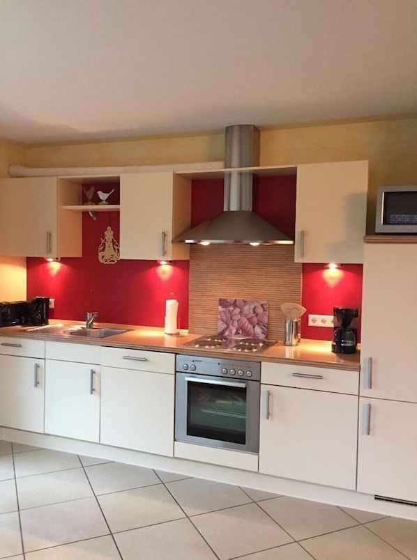 A modern kitchen with white cabinets, a red accent wall, a stainless steel range hood, and a built-in oven.