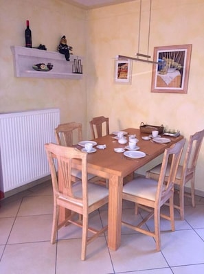 A dining area with a wooden table set for four, surrounded by wooden chairs. There are two shelves above the table, one with a wine bottle and decorative items, and the other with framed pictures.