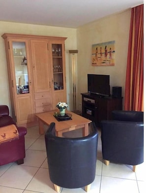 A living room with a wooden cabinet, a television, a table with a flower vase, and two black chairs.
