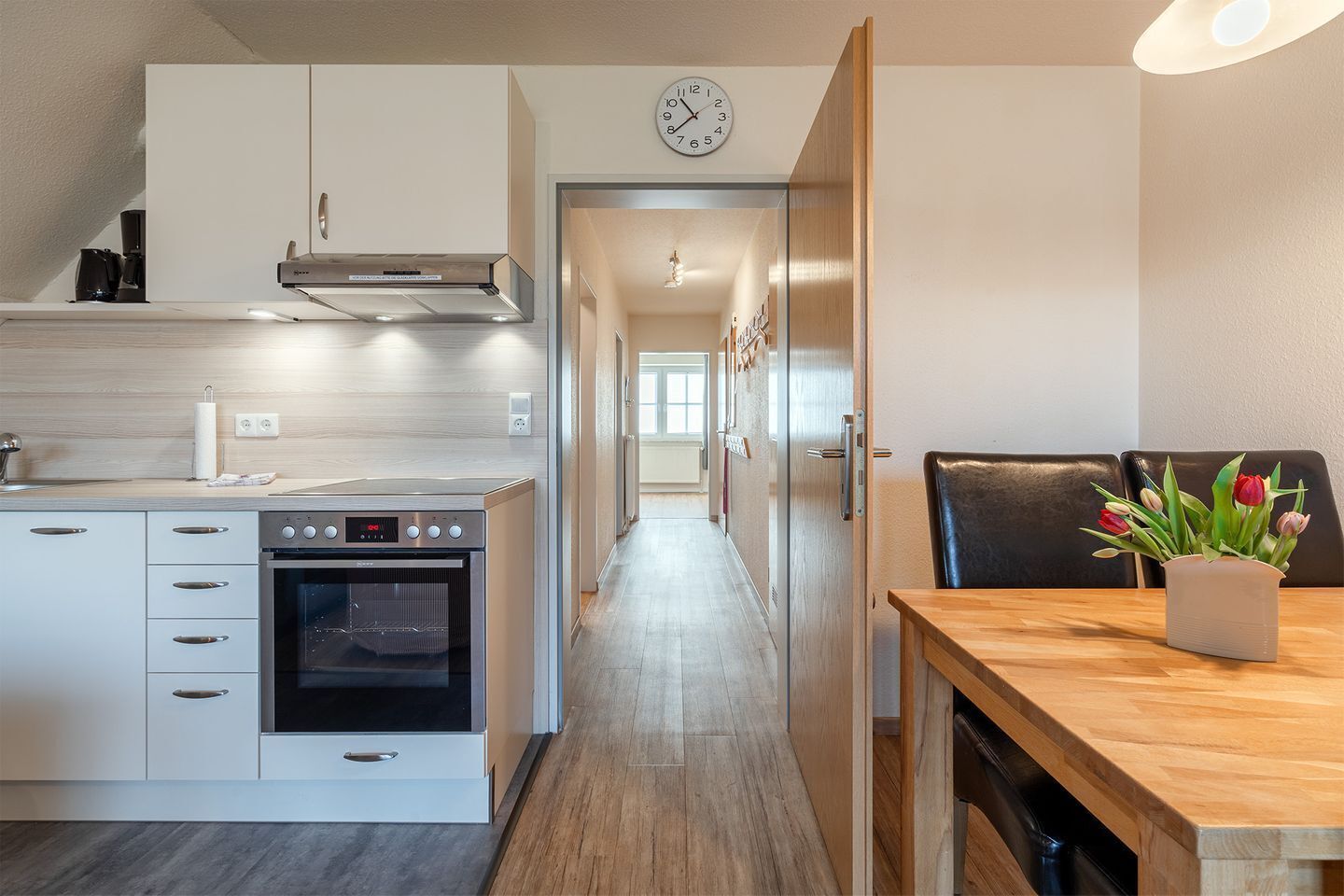 Een moderne keuken met witte kasten, een inbouwoven en een eethoek met een houten tafel en stoelen.