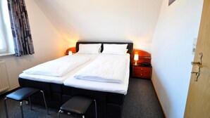 Uma cama de casal com roupa de cama branca, uma mesinha de cabeceira com abajur, um banquinho pequeno e uma janela com cortinas.