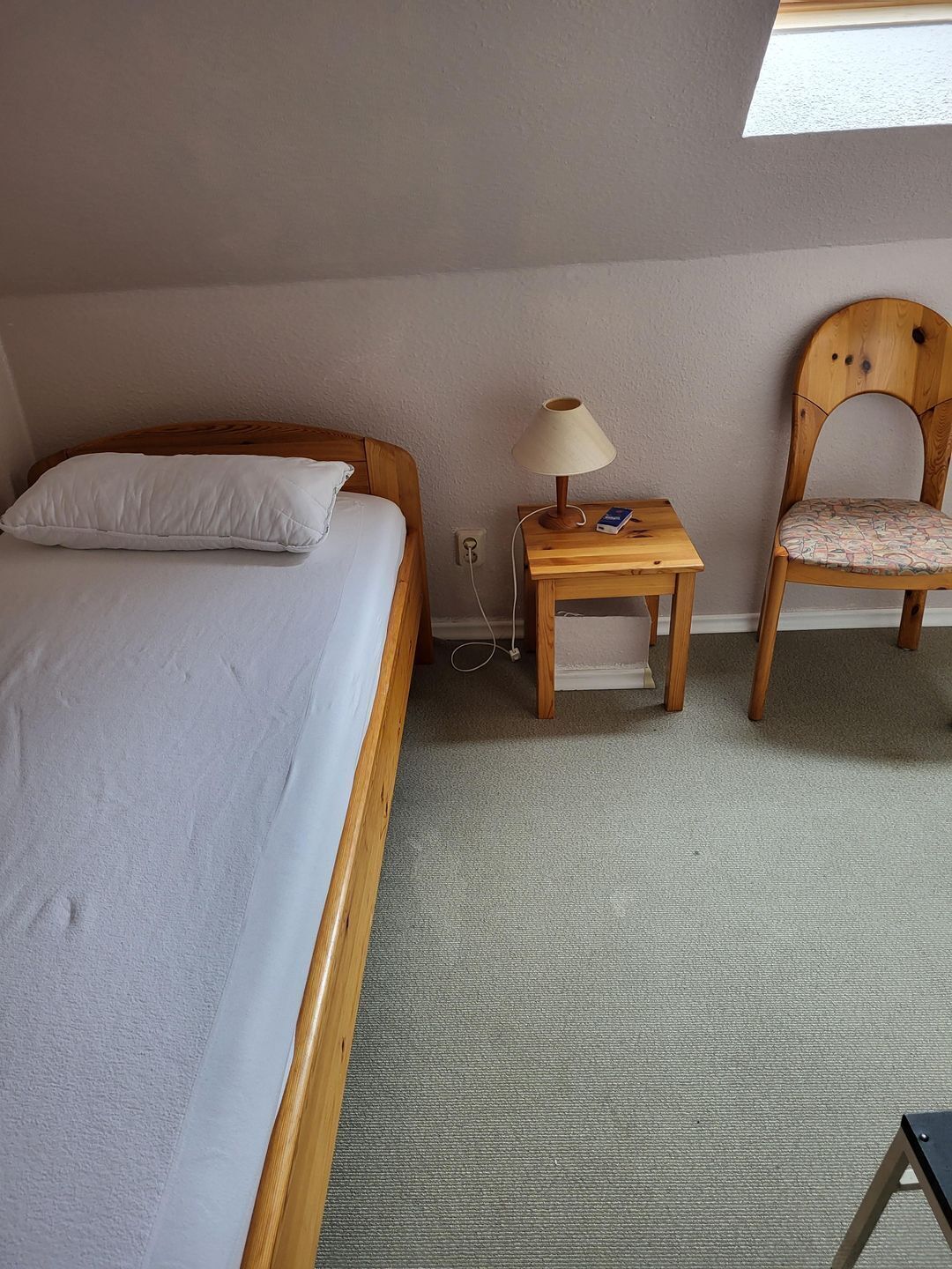 Een slaapkamer met een bed, nachtkastje met lamp, een stoel en een raam.
