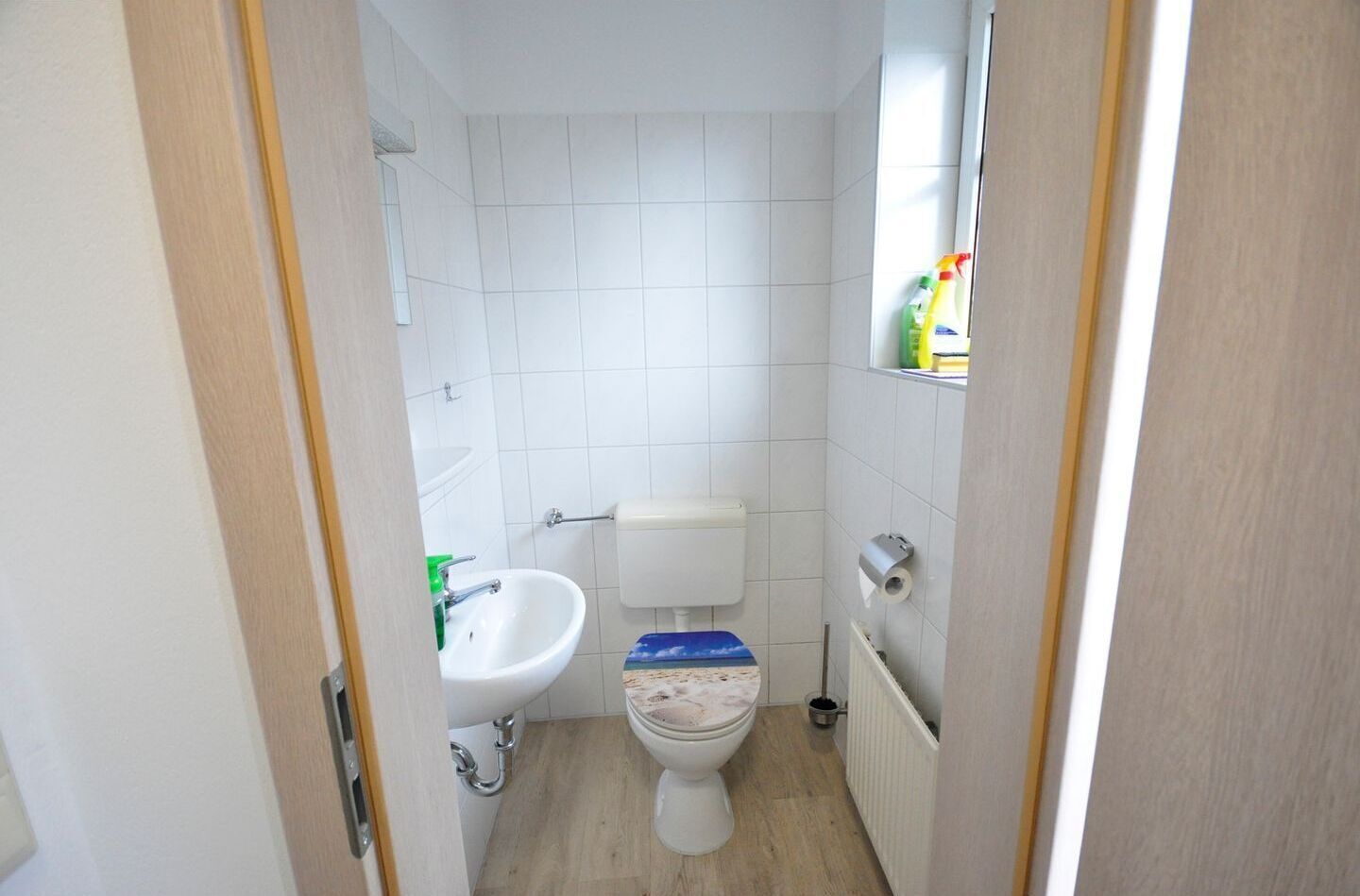 Une petite salle de bain propre, avec des carreaux blancs, un lavabo blanc, des toilettes et une fenêtre donnant sur l’extérieur.