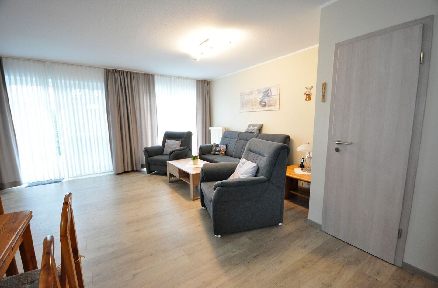 Un salon moderne avec un canapé gris, une table basse en bois et une table à manger avec des chaises. Il y a une porte sur la droite et une fenêtre avec des rideaux sur la gauche.