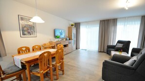 A modern living room with a dining area, a flat-screen TV, and a large window with curtains. - Terraced house Am Haustierpark House 11 M - Am Haustierpark (Werdum)