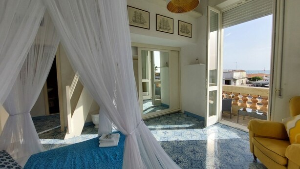 A bedroom with a canopy bed, a balcony with yellow chairs, and a view of the sea. - Imperiale front beach Apartment  (Viareggio)