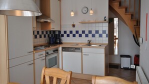 Una cocina compacta equipada con estufa, horno, lavavajillas y cafetera. Se observa una silla de madera y una mesita. En la pared, hay un reloj y un estante con tazas.