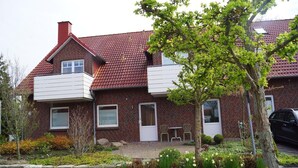 Une maison à deux étages, construite en briques, avec un toit recouvert de tuiles rouges, un balcon et un jardin agrémenté de diverses plantes et fleurs.