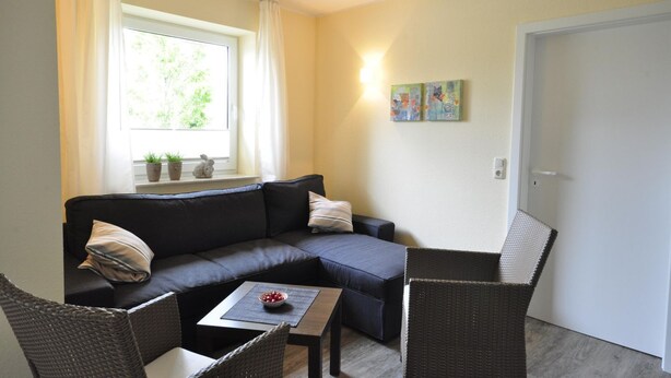 Ein gemütliches Wohnzimmer mit einer Couch, zwei Sesseln, einem kleinen Tisch mit Obstschale und einem Fenster mit Vorhängen.