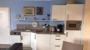 Eine kompakte Küche mit weißen Schränken, einer Mikrowelle und einer Kaffeemaschine. An der Wand hängen zwei gerahmte Bilder und eine kleine Ablage mit Tassen.
