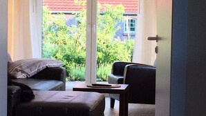 Ein Zimmer mit einer Schiebetür, die auf einen Balkon mit rotem Ziegeldach und viel Grün führt. Im Inneren befindet sich eine dunkelbraune Ledersofa und ein Holztisch.