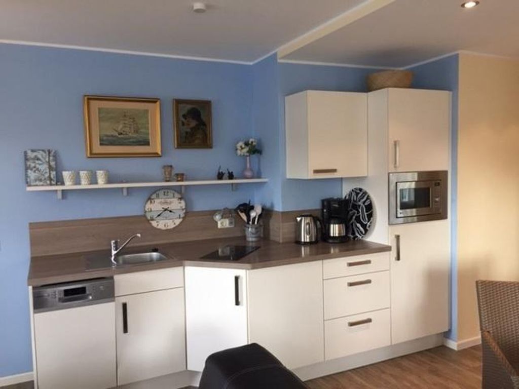 Cocina compacta con armarios blancos, microondas y cafetera. Hay dos cuadros enmarcados en la pared y un reloj.